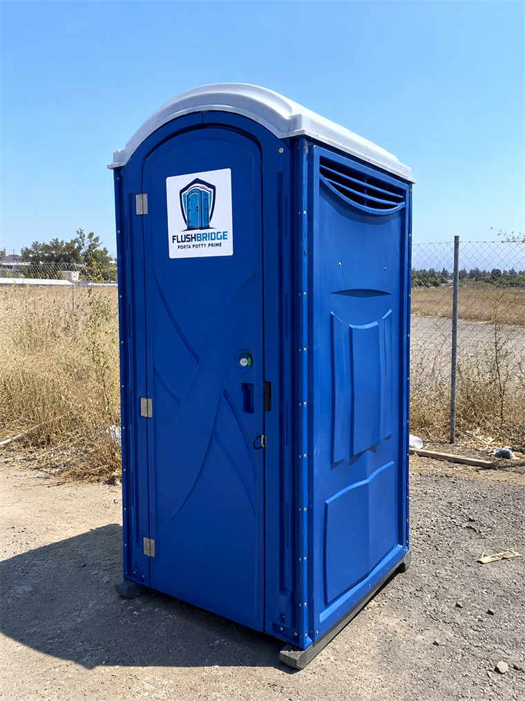 Blue standard portable toilet standing on a gravel lot beside a wire fence