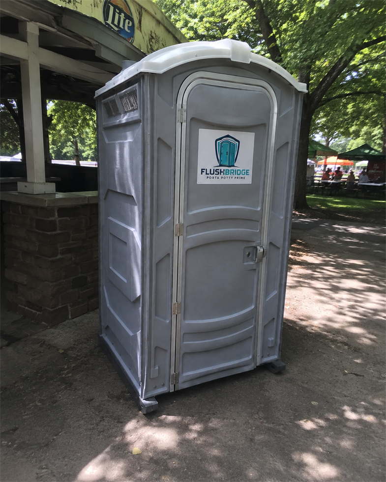 Gray deluxe portable toilet standing in the shade of an open park shelter